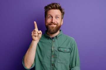 Wall Mural - Photo of an enthusiastic man with a thin beard wearing a casual t-shirt while raising his index finger up against a purple background