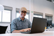 © JoseIMartin - Cheerful Latino man with glasses and flat cap working on a laptop in a bright and spacious office, smiling at the camera.