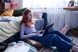 © AnnaStills - Red-haired teen girl sitting on pillow on floor in her bedroom typing on laptop, copy space