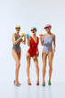 © master1305 - Three beautiful women in colorful swimsuits, swimming caps and sunglasses standing on heels and drinking cocktails against white background. Concept of summer, retro style, fashion