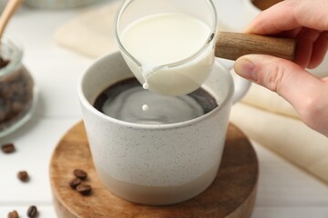 Wall Mural - Woman pouring milk into cup with coffee at white table, closeup