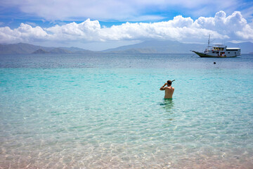Wall Mural - A man wades into the pristine waters off a pink sand beach, with a scenic boat and distant mountains in view.