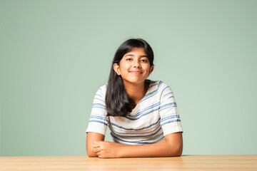 Wall Mural - Indian asian small girl sitting at table against plain green background