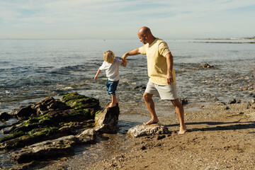 father supports son on rocks in the sea. Shot of father helping his son to jump over the rock on the beach. Young man and little boy having fun at the rocky beach.