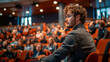 © khonkangrua - A young curly-haired man attentively sitting in a conference auditorium filled with people.