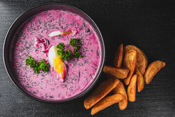 A bowl of beetroot soup with a dollop of cream, garnished with herbs and lemon, served with fried potato wedges on the side