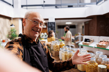 Canvas Print - Older zero waste shop entrepreneur using selfie camera to show his natural pesticides free products online. Senescent supermarket owner promoting sustainable lifestyle, presenting his food stocks