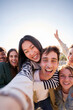 © CarlosBarquero - Vertical. Group of diverse happy young friends taking selfie together looking smiling at camera. Millennial people sharing cheerful community moments posing piggyback with peace gestures raised