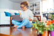 © simona - Attractive adult woman using laptop sitting onthe floor in living room at home in indoor leisure activity surfing the net. Modern people lifestyle and connection. Remote worker in alternative office