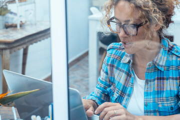 Wall Mural - Smiling woman in checked shirt working on laptop while sitting at desk near window in a laboratory or at home. Vision through the window. Female people with new alternative small business at home