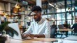 © Scott - A young professional sitting at a desk in a bright office space, typing on a laptop and surrounded by colleagues dressed in business casual attire.