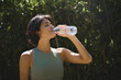 © insta_photos - Active sporty fit young Hispanic healthy woman wearing sportswear holding bottle drinking water hydrating body after doing sport fitness training standing in green nature park outdoor, hydration.