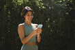© insta_photos - Happy Hispanic fit young woman using mobile phone checking fitness applications standing in green nature park. Online free trainings exercises, sport and health care commercial apps concept.