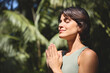 © insta_photos - Calm happy young Hispanic woman holding hands in namaste meditating doing yoga breathing spiritual practice at retreat with eyes closed feeling peace of mind, standing in green nature tropical park.