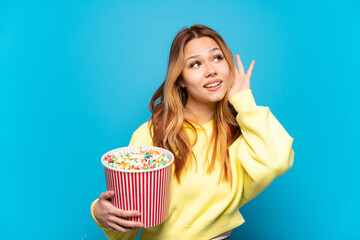 Wall Mural - Teenager girl holding popcorns over isolated blue background listening to something by putting hand on the ear