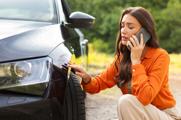 Sad young woman sitting near scratched auto and calling husband for help, having problem with new vehicle