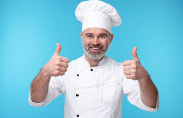 Wall Mural - Happy chef in uniform showing thumbs up on light blue background