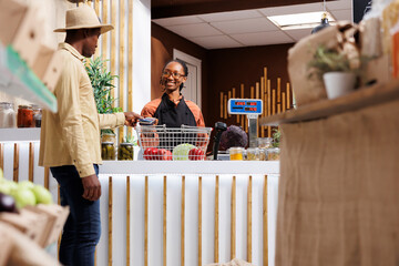 Canvas Print - A young African American man in a hat shops in a contemporary supermarket, browsing items and paying with his credit card. The POS terminal at the checkout counter is operated by the shop owner.