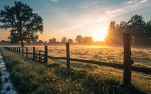 Sunrise Over A Dewy Meadow With A Rustic Wooden Fence And Tree Silhouettes.