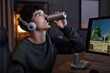 © New Africa - Young man with energy drink and headphones playing video game at wooden desk indoors