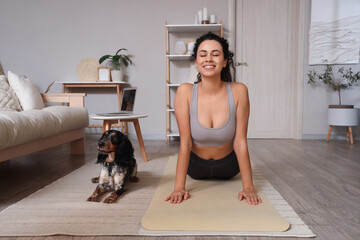 Sporty young African-American woman with cocker spaniel doing yoga on mat at home