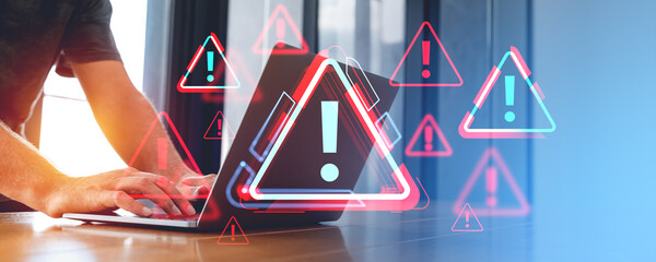 Person typing on a laptop with holographic warning symbols on screen in an office environment, concept of cybersecurity threat