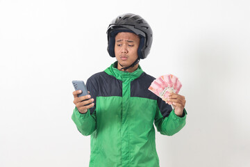 Wall Mural - Portrait of Asian online taxi driver wearing green standing against white background, smiling and looking at camera, holding a bunch of rupiah money