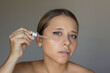 © Марина Демешко - A young caucasian woman applying anti-wrinkle serum to the skin with a dropper on a dark gray background. Nourishing moisturizing cosmetic serum. Cosmetology and beauty concept. Facial care