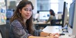 © Summit Art Creations - A young woman sits at her desk and smiles at the camera. She is wearing a blue and black patterned blouse and has long brown hair. There are two computer monitors on her desk and a woman in the