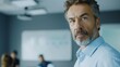 © Z A C - Male actor with gray beard and hair wearing a light blue shirt standing in an office with a whiteboard in the background looking directly at the camera with a serious expression.