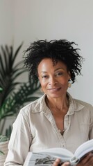 Poster - A woman with curly hair wearing a light-colored blouse smiling and holding a book standing in front of a blurred green plant.