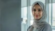 © iuricazac - A woman with a gentle smile wearing a light-colored hijab standing in a modern well-lit room with large windows.