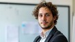 © Z A C - Young man with curly hair and beard wearing a dark suit and light shirt standing in front of a whiteboard with papers attached.