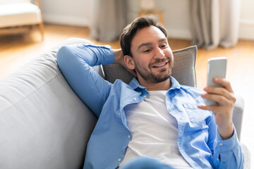 Wall Mural - A man is seated on a couch, engrossed in his phone screen. He appears focused as he scrolls and interacts with the device.