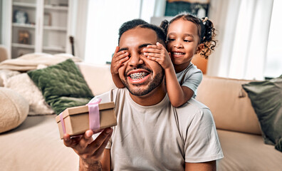 A cute little girl greets her dad by giving him a gift and covers his eyes with her hands to make a surprise.