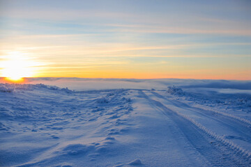 Winter deserted road at sunset, tire ruts on snowy path Picturesque view of Russian countryside Eco tourism tours to Kamchatka, untouched nature Snowdrifts near road, blue sky in rays of orange sun