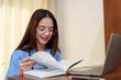 © Ekkasit A Siam - Asian female medical student with long hair, blue scrubs, stethoscope, glasses, smiling while reading a book next to laptop. Bright room with natural light. Concept of education, healthcare, joy.