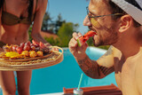 Fototapeta  - Man eating watermelon by a pool and a woman with a tray of fruits and snacks