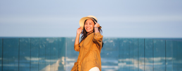 Wall Mural - Portrait image of a young woman with hat, hair blows in the wind, glass terrace with sea and blue sky background