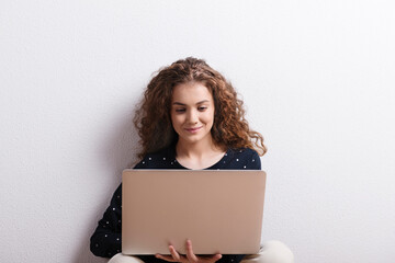 Wall Mural - Portrait of gorgeous teenage girl sitting, typing on notebook. Studio shot, white background with copy space