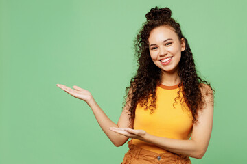 Wall Mural - Young happy woman of African American ethnicity wears yellow tank shirt top point hands arms aside on area mock up isolated on plain pastel light green background studio portrait. Lifestyle concept.