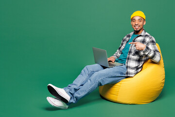 Wall Mural - Full body young man of African American ethnicity he wear shirt blue t-shirt yellow hat sit in bag chair hold use work point on laptop pc computer isolated on plain green background Lifestyle concept