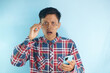 © Lailar Studio - Young Asian man in red shirt showing shocked expression with hands touching glasses frame and holding mobile phone
