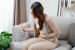 © Suriyawut - The woman with a stomachache is holding her stomach in pain, sitting on the sofa in the living room.