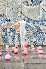 Wall Mural - Young hairless girl ballerina with alopecia in white futuristic suit dancing outdoor and jumps among pink spheres on abstract mosaic Soviet background, symbolizes self expression