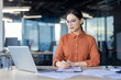 © Liubomir - Asian businesswoman working on her laptop while taking notes in a modern office setting. She looks focused and professional.
