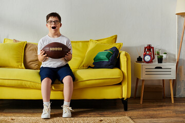 Wall Mural - A adorable boy with Down syndrome holding a football, seated on a bright yellow couch.