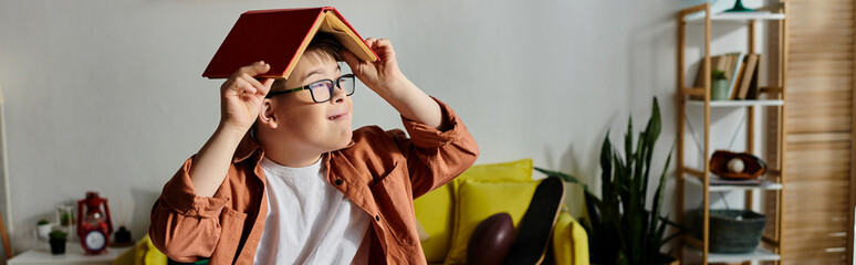 Wall Mural - Little boy with Down syndrome holds a book over his head while sitting on a couch.