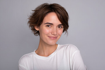 Close up portrait of gorgeous young woman with healthy skin and modern hairstyle looking on camera on grey wall background