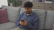 © Krakenimages.com - A young man with a beard engaged on his smartphone while sitting on a couch indoors.